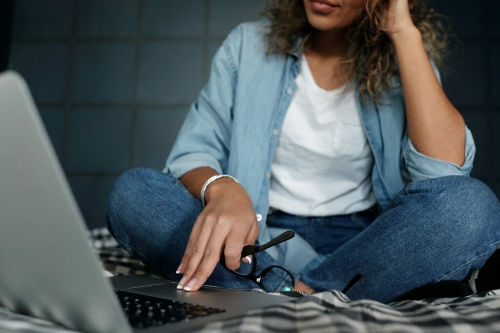 Woman sitting cross-legged on bed looking at laptop while holding glasses