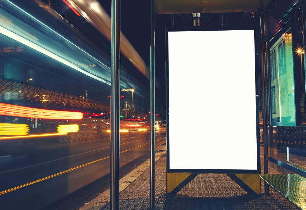 Illuminated blank billboard with copy space for your text message or content, advertising mockup banner of bus station, public information board with blurred vehicles in high speed in night city